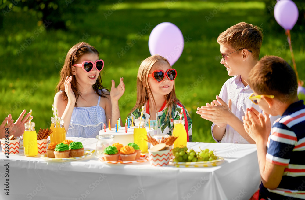 holidays, childhood and celebration concept - happy kids with candles on cake sitting at table at summer garden party and applauding to birthday girl holidays, childhood and celebration concept - happy kids with candles on cake sitting at table at summer garden party and applauding to birthday girl