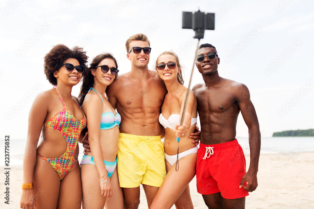 friendship, summer holidays and people concept - group of happy friends taking picture by smartphone on selfie stick on beach