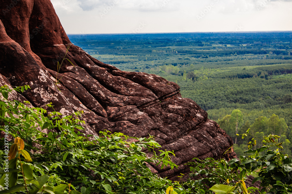 A red rock cliff side high above the ground. Rain forest and deep skies make up the background.