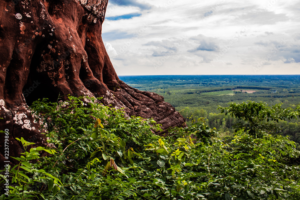 A red rock cliff side high above the ground. Rain forest and deep skies make up the background.