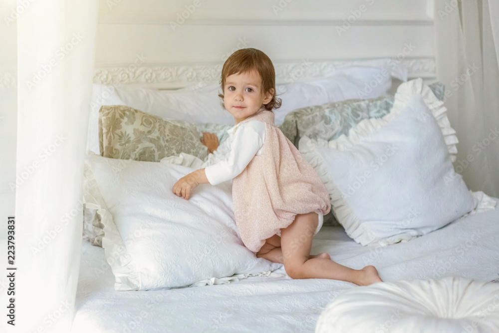 Funny and cute brunette little smiling girl playing jumping on bed in light bedroom. White interior with big bed. Childhood, preschool, youth, relax concept