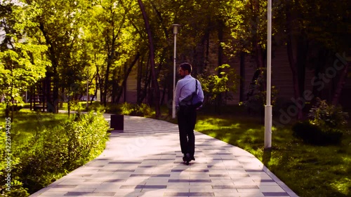 Dolly shot. A man in a business suit riding a kick scooter in a park then he taking scooter and going up stairs. Rear view