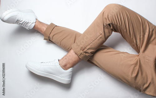 Fragment of male legs in beige trousers and white sneakers sits on a white background. Top view. Relaxation.
