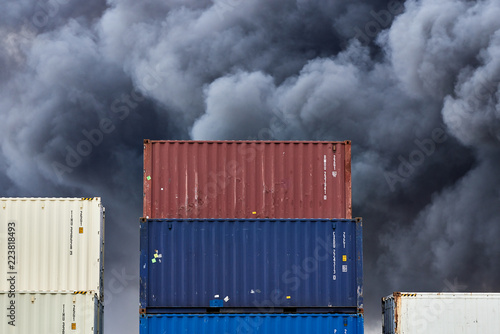 Shipping containers stacked in storage with plumes of black toxic smoke from a fire behind.