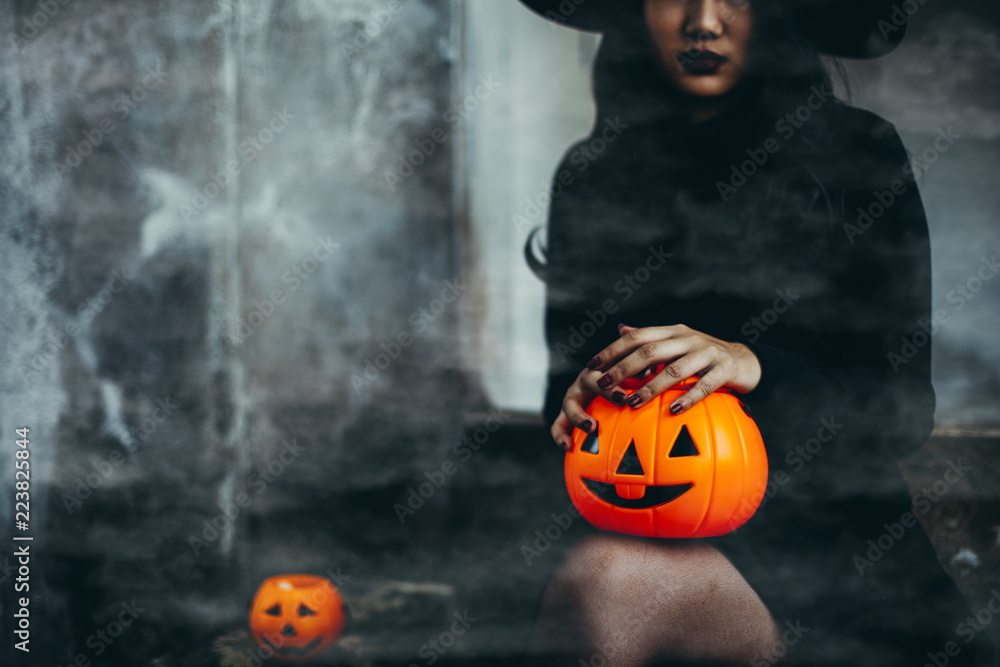 Fototapeta premium Young woman in black dress with witch hat and orange pumpkin in hand is on a black background, Halloween concept. Horror theme.