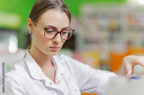 A pretty graceful dark-haired lady with glasses, dressed in a white coat, carefully examies the goods on the shelf in the pharmacy.