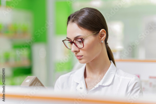 A pretty graceful dark-haired lady with glasses, dressed in a white coat, carefully examies the goods on the shelf in the pharmacy. Girl's profile is shown.