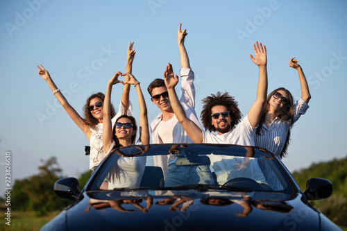 Stylish young girls and guys in sunglasses are sitting in a black cabriolet on the road holding their hands up and smiling on a sunny day.