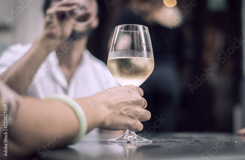 Detail on a woman hand holding a white wine glass, a person drinking in the background