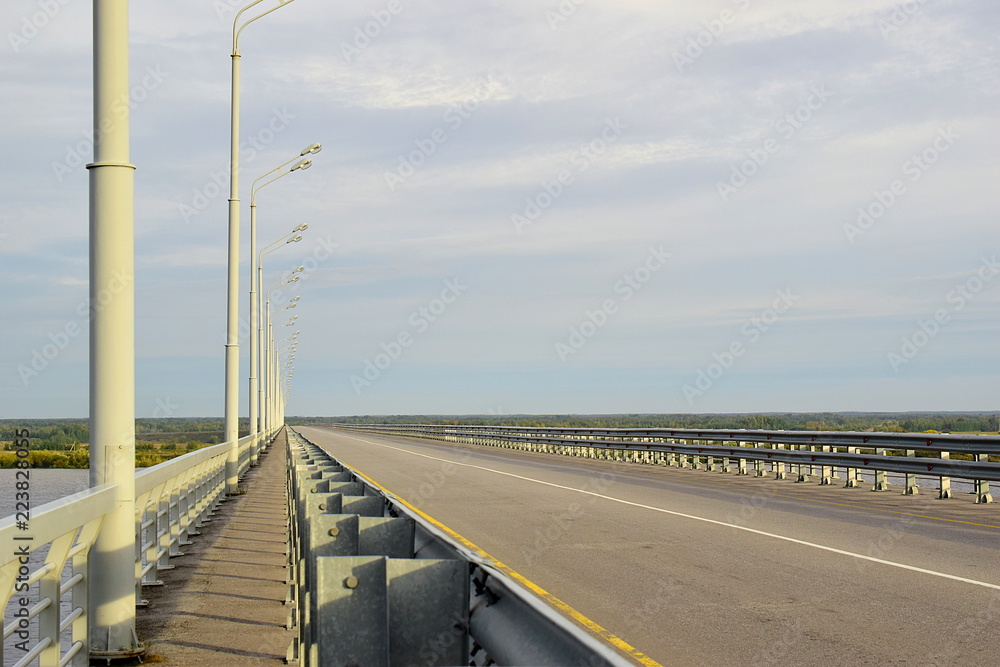 Road bridge over the river. Asphalt pavement, fences and lampposts, erased road markings. Cloudy weather. Copy space.
