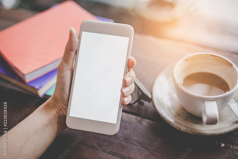 Mockup portrait of a woman hand holding a  cell phone with blank screen and a cup of coffee in cafe.