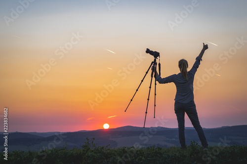 Female photographer holding a tripod with camera and looking at sunset. Woman enjoying spending time with camera in nature.