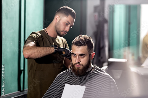 Handsome young man with beard sits at a barber shop. Barber shaves hairs at the side.
