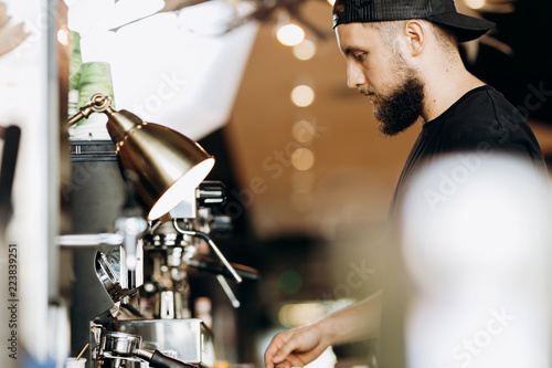 A stylish young man with beard,wearing casual clothes,cooks coffee in a coffee machine in a modern coffee shop.