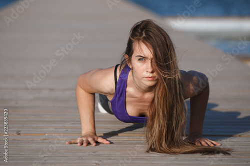 Beautiful young girl with long hair dressed in sports clothes is doing push ups on the wooden pier on the summer day