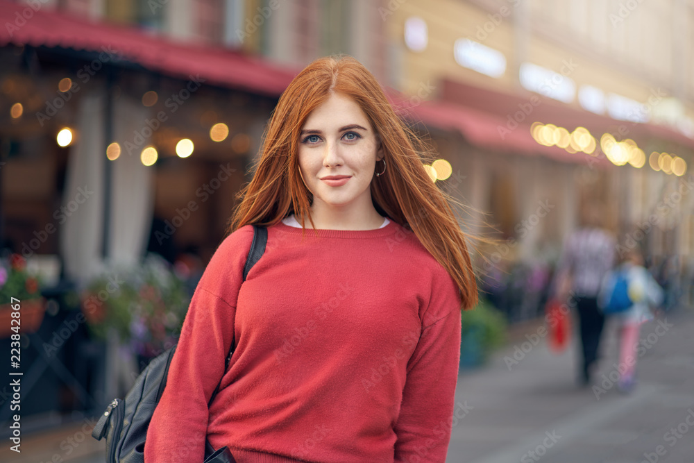 A spectacular red-haired girl with backpack on her shoulder poses for a portrait against the background of a fair street. Positive emotions, a walk through the streets of the city and lifestyle A spectacular red-haired girl with backpack on her shoulder poses for a portrait against the background of a fair street. Positive emotions, a walk through the streets of the city and lifestyle