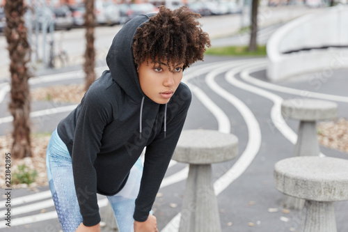 Tired black female runner or sportswoman dressed in casual sweatshirt with hoodie, cathces breath, keeps hands on knees, poses against road or street blurred background. Urban style concept. photo