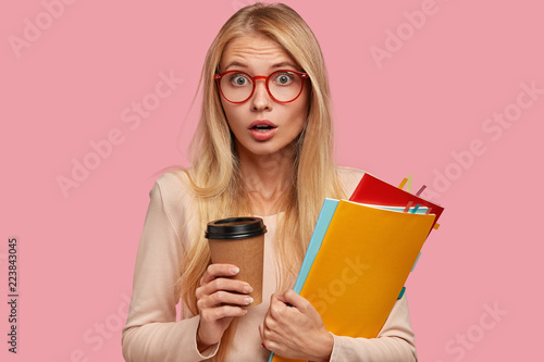 Surprised wondered blonde Caucasian woman comes on training lesson, being shocked to have unexpected task, holds takeaway coffee, wears round spectacles, drinks takeaway coffee, carries books