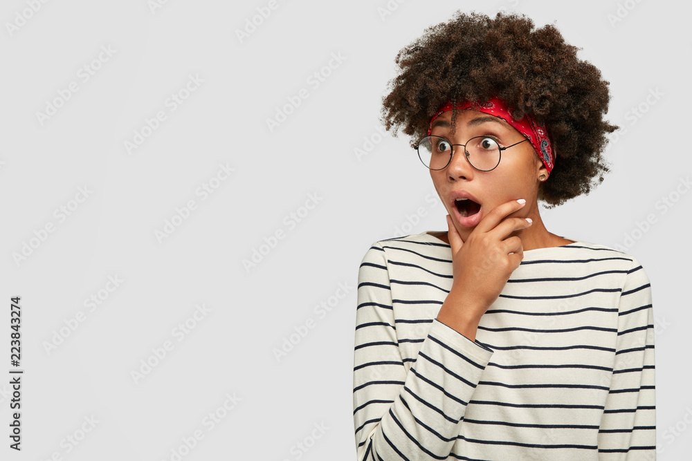Surprised dark skinned woman keeps jaw dropped, holds chin, looks with ...