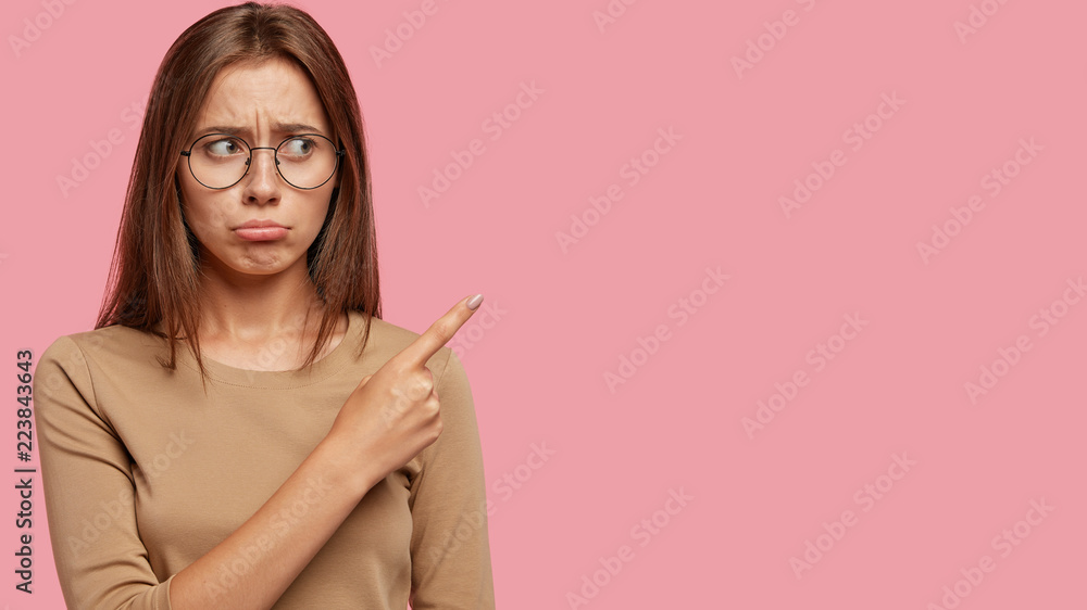 Studio shot of displeased woman purses lips, points with index finger at upper right corner, has unhappy expression, dressed in casual sweater, isolated over pink background. Advertising concept Studio shot of displeased woman purses lips, points with index finger at upper right corner, has unhappy expression, dressed in casual sweater, isolated over pink background. Advertising concept