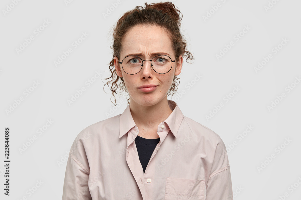 Studio shot of dissatisfied freckled girl has gloomy expression, looks with bewilderment, has freckled face, dislikes something, dressed in fashionable shirt, isolated over white background.