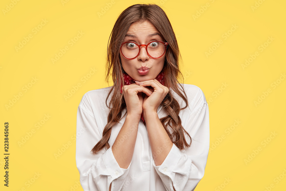 European teenager has amazed look at camera, keeps hands under chin, wears spectacles, white shirt, bandana poses over yellow background, pouts lips, has surprised facial expression. Youth concept European teenager has amazed look at camera, keeps hands under chin, wears spectacles, white shirt, bandana poses over yellow background, pouts lips, has surprised facial expression. Youth concept