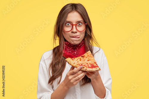 Pretty woman with appealing appearance, licks lips, holds slice of tasty pizza, being hungry, wants to eat, enjoys taste, has good appetite, isolated over yellow studio wall. People and snack concept