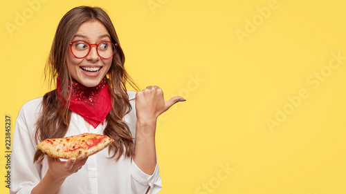 Fastfood and meal concept. Cheerful young girl smiles pleasantly, eats tasty slice of pizza, being in good mood, shows place she bought it, advertises pizzeria, isolated over yellow background.