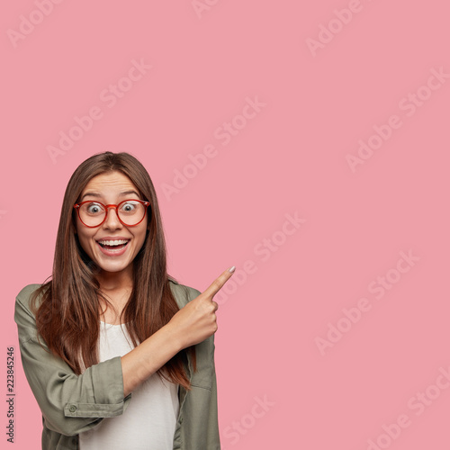 Excited young European woman being under positive impression, points right, sees something incredible, has broad smile, wears round spectacles, poses alone against pink background. Its hilarious!