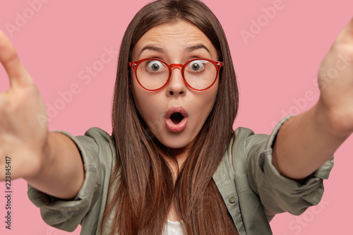 Stupefied Caucasian woman with surprised terrified expression, stretches hands for making selfie, opens mouth from wonder, poses over pink background. Brunette girl in stupor models at studio photo
