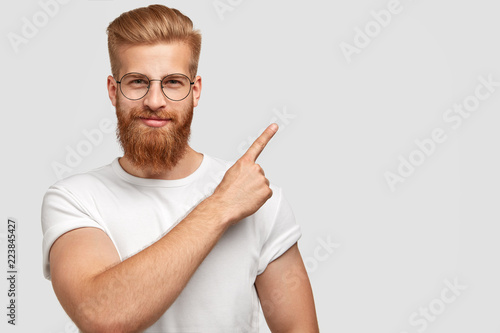 Studio shot of ginger hipster with thick beard, trendy haircut, has serious expression, points with index finger at upper right corner, dressed in white t shirt shows free space for your advertisement photo