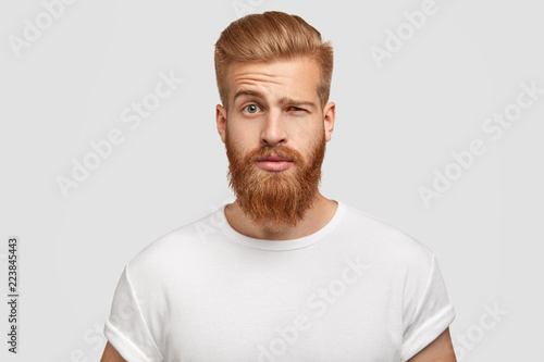 Fotografie, Tablou Bewildered man with thick ginger beard, raises eyebrows, reacts on fake news from friend, looks directly at camera, dressed in casual t shirt, isolated over white background