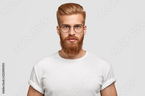 Horizontal shot of attractive hipster looks seriously directly at camera, being self assured, wears big round spectacles, white t shirt in tone with background. Masculinity and style concept © wayhome.studio 