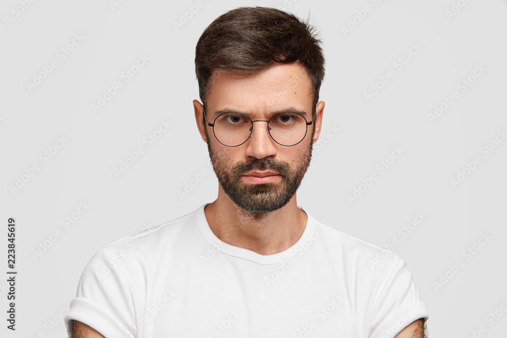Close up shot of confident strict businessman has thick bristle, looks with gloomy expression, frowns face, wears glasses and casual t shirt, expresses negative feeling, isolated over white background Close up shot of confident strict businessman has thick bristle, looks with gloomy expression, frowns face, wears glasses and casual t shirt, expresses negative feeling, isolated over white background