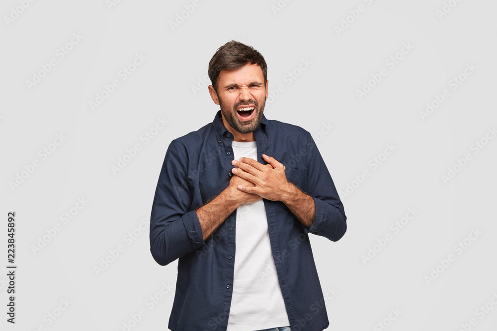 Attractive hipster with positive expression laughs positively, keeps both palms on chest, dressed in blue shirt, poses over white background. Man hears something funny. Good emotions concept Attractive hipster with positive expression laughs positively, keeps both palms on chest, dressed in blue shirt, poses over white background. Man hears something funny. Good emotions concept