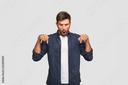 Emotional stupefied young European male adult notices something stunning on floor, points with index fingers on floor, opens mouth from wonder, dressed casually, stands against white background