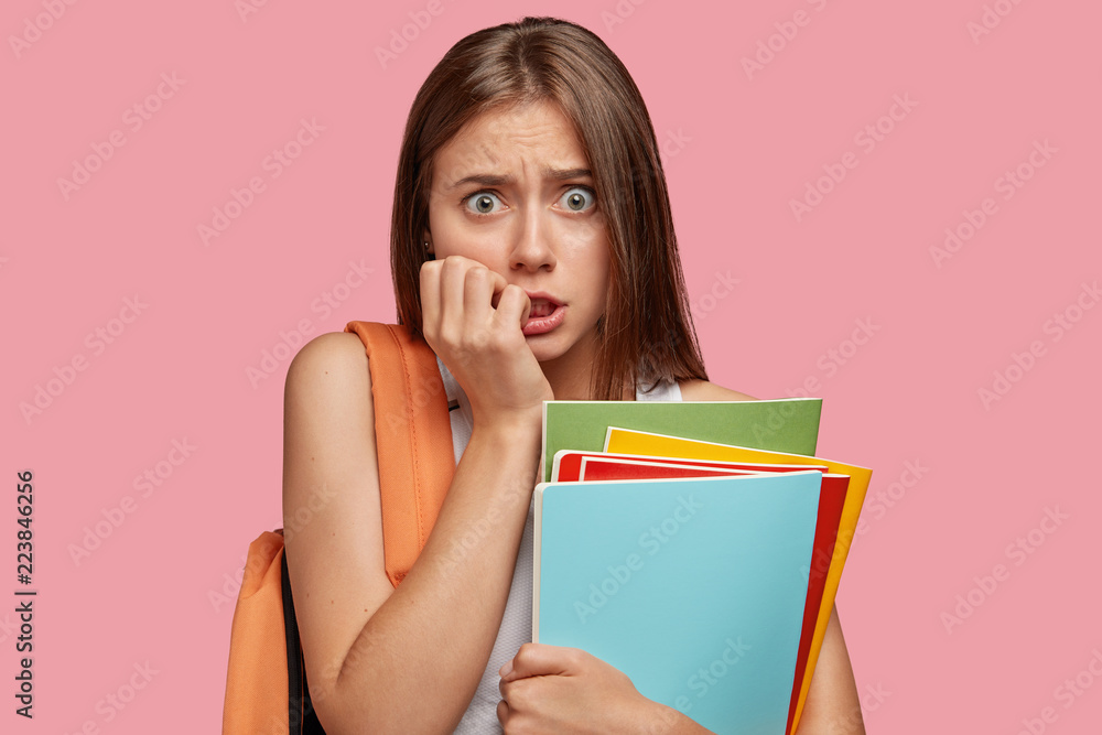 Neurotic puzzled student girl bites finger nails, looks anxiously at camera, worries before answering during class, not sure in her knowledge, stands against pink background. Frustrated schoolgirl Neurotic puzzled student girl bites finger nails, looks anxiously at camera, worries before answering during class, not sure in her knowledge, stands against pink background. Frustrated schoolgirl