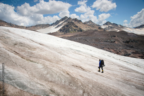 A mountaineer with a backpack walks in crampons walking along a dusty glacier with sidewalks in the hands between cracks in the mountain