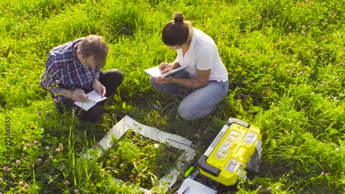 Crane shot. Two ecologist examining plants on the meadow. Woman taking protective gloves from toolbox. Field work. High angle view