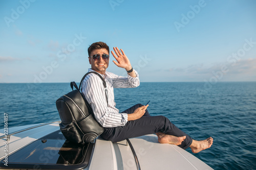 Positive caucasian freelancer with backpack over his shoulder spending resttime at sea journey, smiling at camera, enjoys sunset from yacht. photo