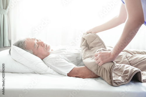 Senior man sleeping in bed and nurse put blanket on him.