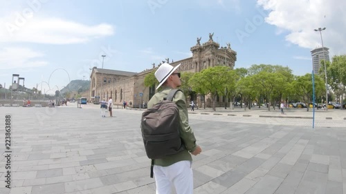 A young man, tourist standing with a backpack and in a hat and takes photos by smartphone near the Columbus Monument, port area on  