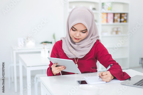 Young asian muslim woman using tablet and taking notes in notebook in a white cafe.