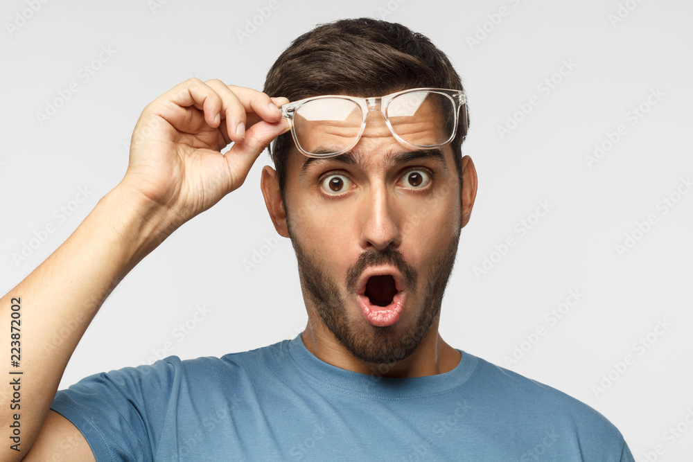 Young man in blue t-shirt shouting OMG with open mouth, surprised by low price and sales, holding transparent glasses Young man in blue t-shirt shouting OMG with open mouth, surprised by low price and sales, holding transparent glasses