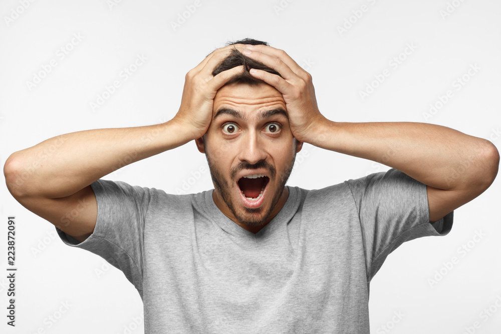 Emotional young man with strong expression of fear, round eyes, open mouth and hands pressed to top of head, showing astonishment and shock, isolated on gray background Emotional young man with strong expression of fear, round eyes, open mouth and hands pressed to top of head, showing astonishment and shock, isolated on gray background