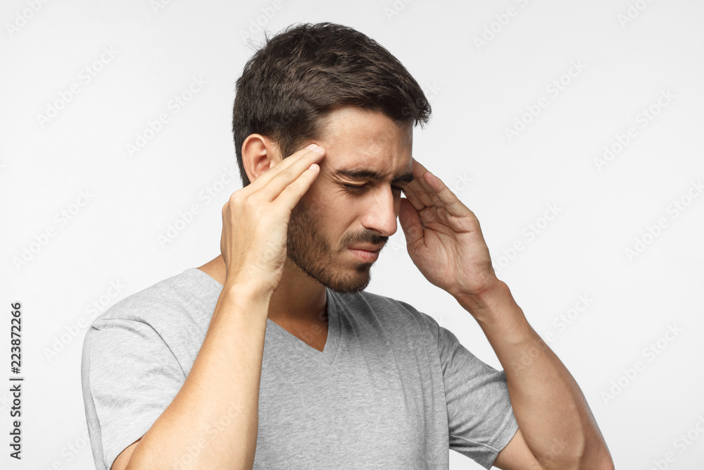 Closeup of young man isolated on gray background in casual grey t-shirt, touching temples with fingers as if suffering from severe migraine Closeup of young man isolated on gray background in casual grey t-shirt, touching temples with fingers as if suffering from severe migraine