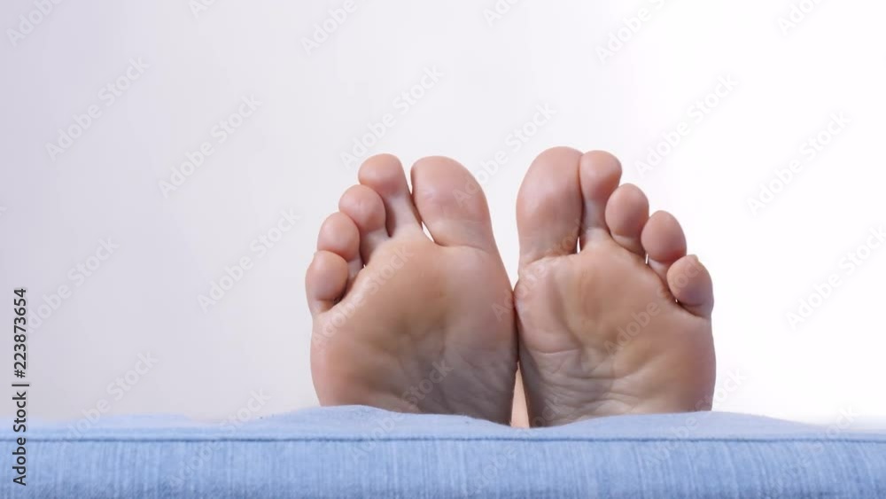 Female resting on sofa showing bare foot. Human feet moving on bed in slow motion, white grey wall background copy space text area Female resting on sofa showing bare foot. Human feet moving on bed in slow motion, white grey wall background copy space text area