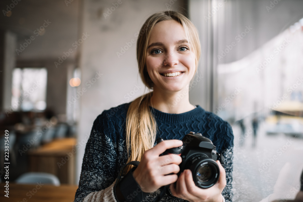 Professional photographer holding digital camera. Smiling hipster woman with beautiful face and blue eyes standing in loft cafe.  - obrazy, fototapety, plakaty 