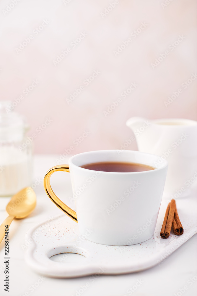 White cup of tea, cinnamon and milk jug on serving plate, golden spoon and sugar on white marble table top. Feminine rose background with copy space. Narrow depth of field.