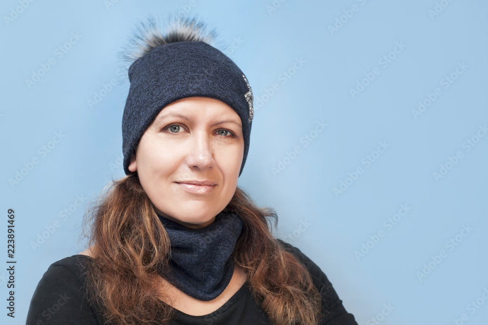 A beautiful middle-aged woman in a warm hat and a scarf on her neck smiles on a blue background A beautiful middle-aged woman in a warm hat and a scarf on her neck smiles on a blue background